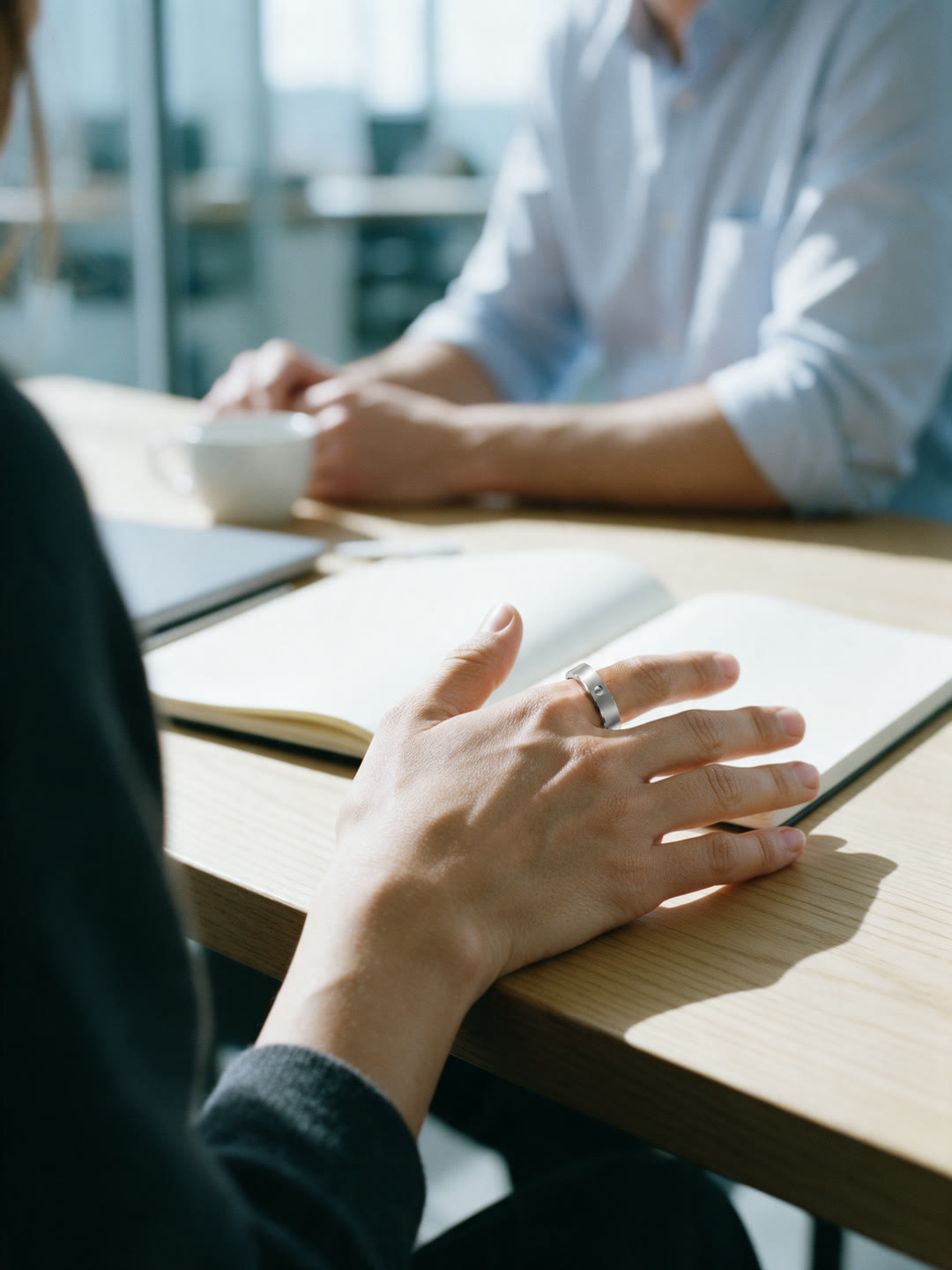 Generated image: over a woman's right shoulder at a meeting-room table, her right hand resting flat beside a blank notebook, the ring on her index finger beside the thumb, a colleague in a pale blue shirt soft across the table