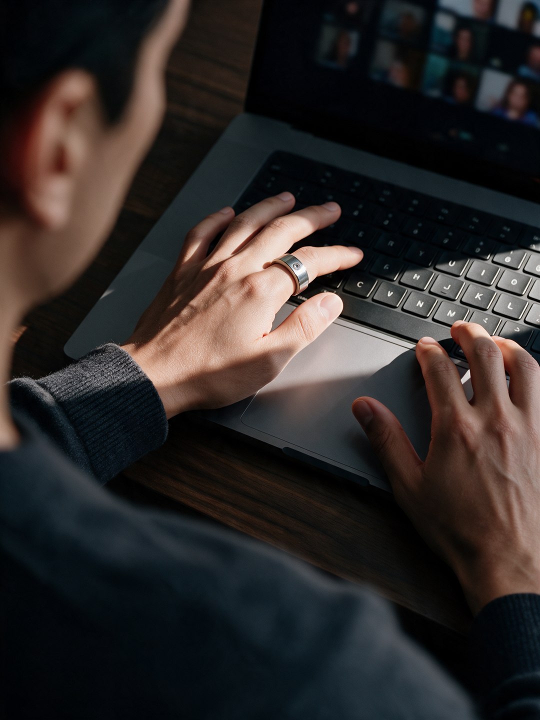 Generated image: over a man's shoulder, both hands typing on a laptop in a strip of sun, a video call on the screen, the ring on his left index finger